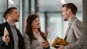 Three professionals having a conversation in a modern office hallway.